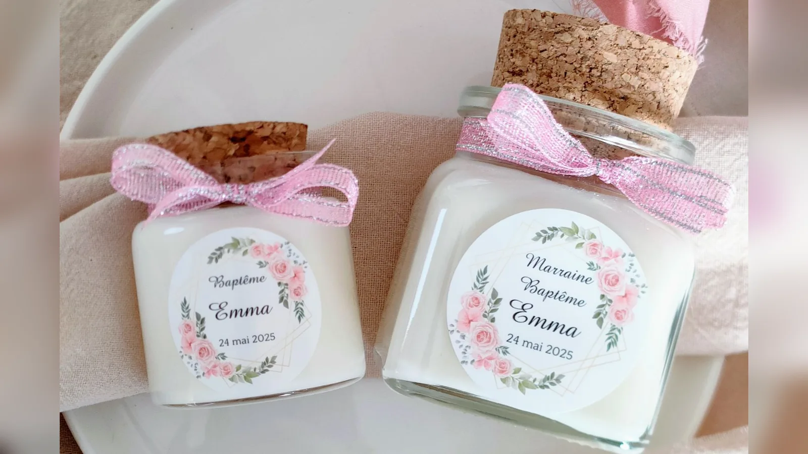 Deux bougies de baptême personnalisées en pot de verre, fermées par un bouchon en liège et un ruban rose, avec les étiquettes « Baptême Emma » et « Marraine Baptême Emma ».